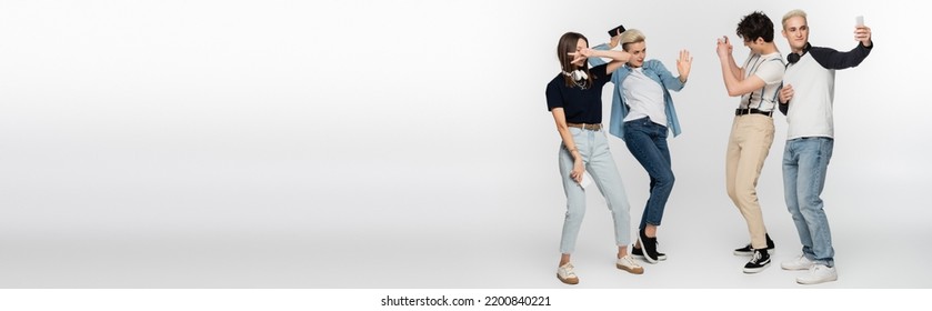 Young Woman Showing Victory Sign And Dancing Near Friends Taking Photo On Smartphones On Grey Background, Banner