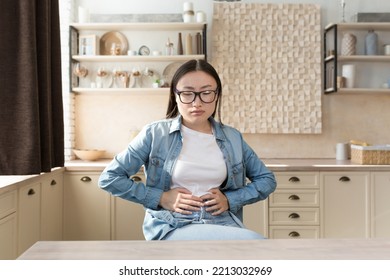 Young Woman Sick Alone At Home Having Severe Stomach Pain, Asian Woman Holding Her Stomach With Hands Sad Sitting On Sofa In Living Room