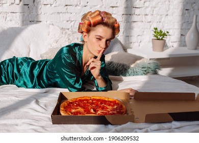 young woman in silk pajama showing silence gesture with pizza pepperoni in bed