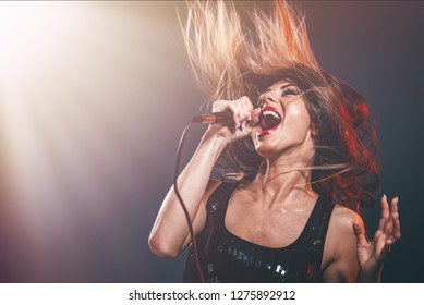 A young woman singer with tousled long hair holding a microphone and sing with a wide open mouth.