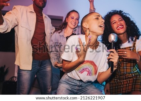 Young woman singing her heart out on karaoke night. Vibrant young woman entertaining her friends with a microphone at a house party. Group of happy young people having a good time on the weekend.