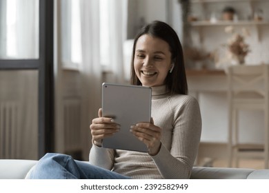 Young woman sit on sofa wear wireless earphones looks at digital tablet screen smile watch funny videos spend weekend leisure at home enjoy pleasant conversation use video call application on device.