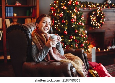 Young Woman Sits in an Armchair at Home Against Background of Christmas Tree and Fireplace. Hold Cup of Hot Drink in Hands. Merry Christmas and Happy New Year greeting card