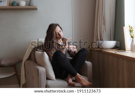 A young woman sits in a comfortable chair by the window in the house and drinks tea. Home interior design. Freelancer