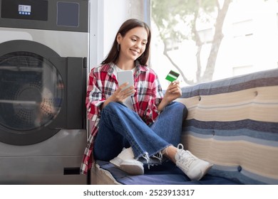 A young woman sits comfortably on a couch in a laundromat, smiling while checking her smartphone and holding a green card. Sunlight brightens the space, creating a relaxed atmosphere. - Powered by Shutterstock