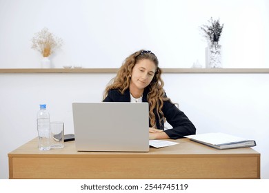 A young woman sits at a contemporary wooden desk, engaged in work on her laptop. Papers and a water bottle are placed nearby, creating a tidy workspace in a bright, minimalist office environment. - Powered by Shutterstock