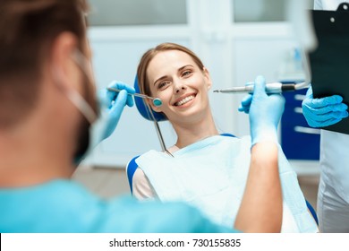 A young woman sits in a dental chair and smiles. Doctors bowed over her. They are going to treat her teeth. Happy patient and dentist concept.
