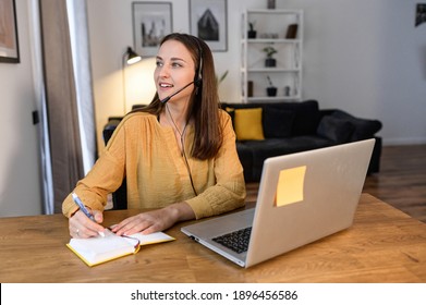 A Young Woman Sits At The Desk, Using Hands Free Headset And Laptop For Remote Work, Talking Online And Taking Notes. Female Call Center Operator Works Remotely