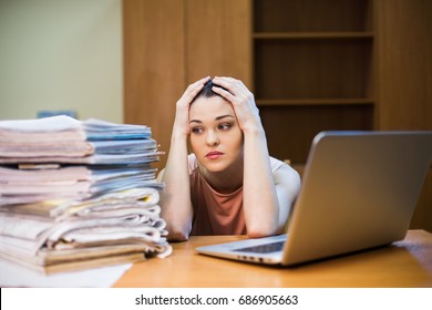 A Young Woman Sits In Front Of A Pile Of Papers And A Computer Holding Her Head