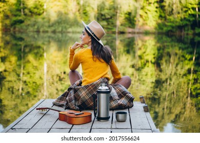 Young woman sits with a guitar on a bridge on a lake with an autumn landscape. Toning.