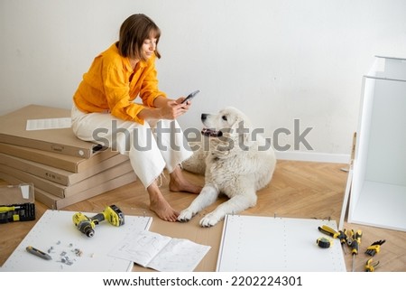 Young woman sits with her cute dog, using phone during repairing at new apartment. Assembling furniture by herself. DIY concept
