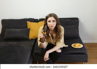 A Young Woman Sits At Home On The Couch And Watches TV Alone. She Drinks Beer And Eats Chips.