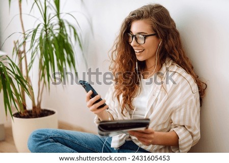 Young woman sits at home, using smartphone, learning, reading book. Resting, education, technology concept.