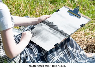 a young woman sits on the beach and draws a landscape on canvas with charcoal.