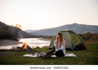 Young woman sits on a blanket on the grass near green tourist tent and bonfire. Girl drinks hot tea or coffee from thermos cup on the background of river and mountains. Resting and hiking concept - Powered by Shutterstock