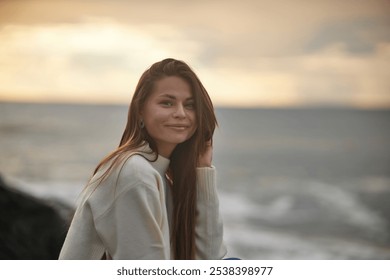 A young woman sits on a rocky shore, gazing into the distance with a calm expression. Dressed casually in jeans and a light sweater, she enjoys the serene ocean backdrop at sunset. - Powered by Shutterstock