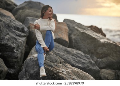 A young woman sits on a rocky shore, gazing into the distance with a calm expression. Dressed casually in jeans and a light sweater, she enjoys the serene ocean backdrop at sunset. - Powered by Shutterstock