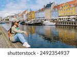 A young woman sits on a wooden pier overlooking the canals of Copenhagen, her gaze fixed on the water. The colorful buildings and boats reflect in the calm water, creating a picturesque scene.