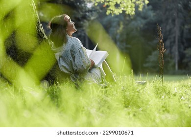 A young woman sits in a park leaning against a tree, reading a book in spring or summer, relaxed and happy, enjoying the sun. - Powered by Shutterstock