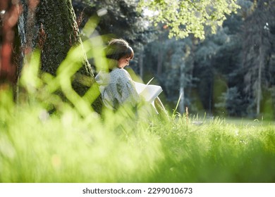 A young woman sits in a park leaning against a tree, reading a book in spring or summer, relaxed and happy, enjoying the sun. - Powered by Shutterstock