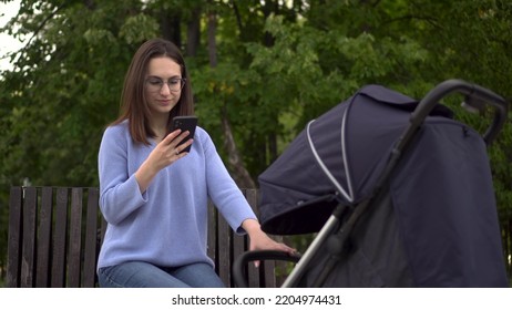 A Young Woman Sits With A Phone In Her Hands And Shakes The Stroller. Modern Mom Is Texting In A Smartphone In The Park.