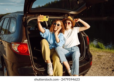 Young woman sits in the trunk of a car and takes selfie. Stylish models on a car trip. The concept of technology, blogging, travel. - Powered by Shutterstock