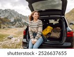 Young woman sits in the trunk of her car, parked on the side of a winding road that cuts through the rocky terrain. Road trips. Autumn landscape