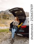 Young woman sits in the trunk of her car, parked on the side of a winding road that cuts through the rocky terrain. Road trips. Autumn landscape