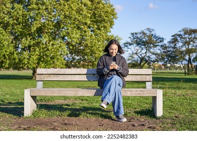 Young woman sitting alone on bench in park, using mobile phone, looking at screen, texting, sending messages in app chat. - Powered by Shutterstock