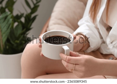 Young woman sitting in armchair and holding cup of delicious coffee