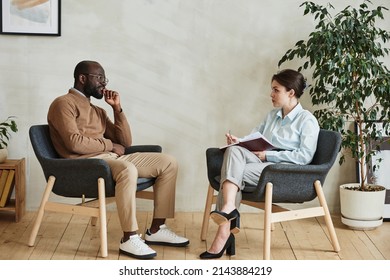 Young Woman Sitting In Armchair And Making Notes In Diary While Giving Individual Psychotherapy To Black Man