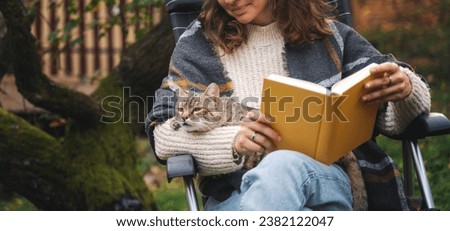 Young woman sitting in the autumn garden with a pet gray cat in her hands reading a book