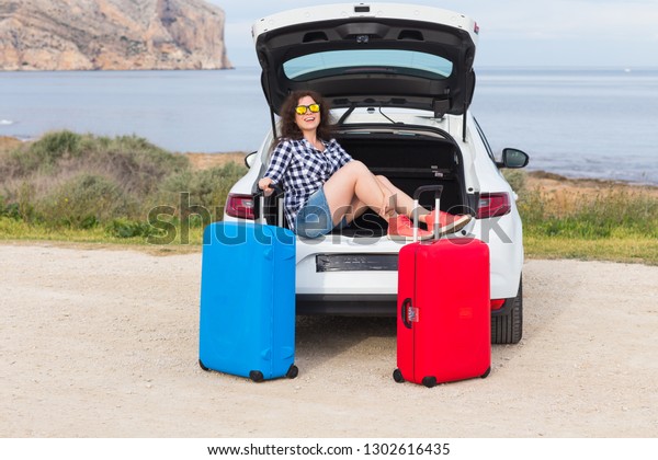 Young woman sitting in back of car smiling and\
getting ready to go. Girl sitting in the open trunk of a car.\
Summer road trip