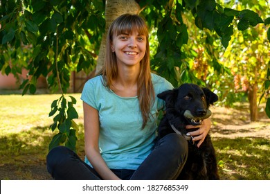 Young Woman Sitting In Backyard On Grass With Beautiful Black Dog.