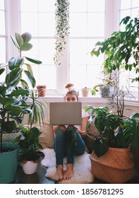 Young woman is sitting by a large window with lots of tropical houseplants using laptop computer