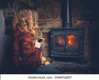 A Young Woman Is Sitting By A Log Burner And Is Using Her Smart Phone
