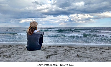 Young woman sitting by the sea with stormy sky, looks at the horizon thoughtfully