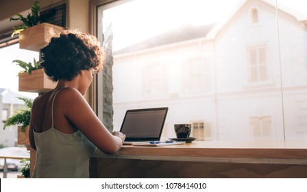 Young Woman Sitting At Cafe Table And Working On Laptop. African Female At Coffee Shop Using Laptop Computer.