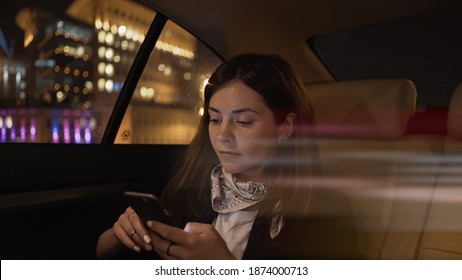 Young woman sitting in car backseat with phone in hands, city lights on background. Woman passenger in black jacket and neck scarf alone with phone, in car late at night
