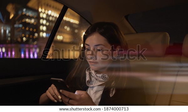 Young woman sitting in\
car backseat with phone in hands, city lights on background. Woman\
passenger in black jacket and neck scarf alone with phone, in car\
late at night