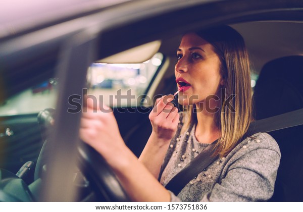 Young woman sitting in\
the car on the driver\'s seat putting the make up on her lips using\
brush lipstick looking to the mirror while waiting on the parking\
at night