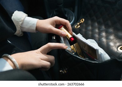 Young Woman Sitting In Car And Taking Lipstick Out Of Handbag