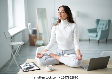 Young Woman Sitting With Closed Eyes Desk Next To Laptop In Padmasana Yoga Pose Home Office.