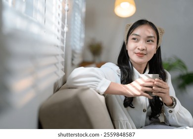 Young woman sitting comfortably on a sofa, holding a warm mug and gazing thoughtfully into the distance, enjoying a peaceful moment in soft sunlight streaming through window blinds - Powered by Shutterstock