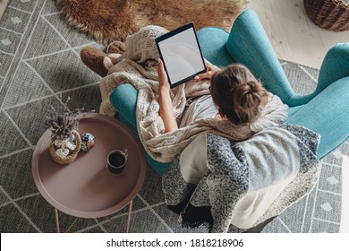 Young woman sitting in cozy armchair, with warm blanket, using tablet