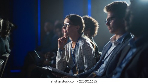 Young Woman Sitting in a Crowded Audience at a Business Conference. Female Attendee Writing Down Important Bullet Points into a Notebook. Keynote Speech in Auditorium with Successful Businesspeople. - Powered by Shutterstock