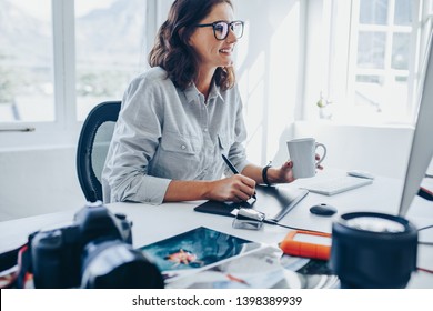 Young woman sitting at desk editing images on computer. Female photographer retouching photos in office using graphic tablet and digital pen. - Powered by Shutterstock