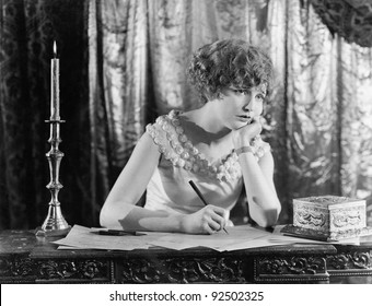 Young Woman Sitting At A Desk With A Pen In Hand, Looking Sad While Writing A Letter