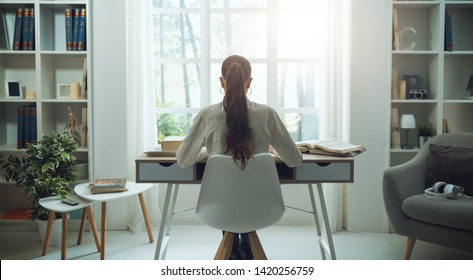 Young woman sitting at desk and studying at home, she is reading books, back view