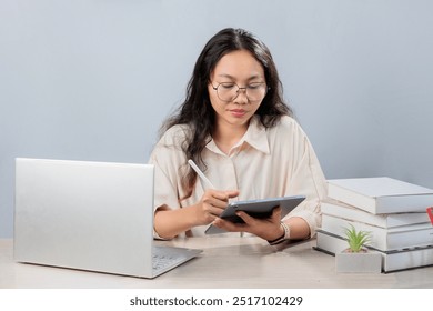 young woman is sitting at a desk working with a laptop and tablet, looking at the tablet. She is wearing glasses and a light-colored blouse. On the desk are stacked books on one side and colorful - Powered by Shutterstock